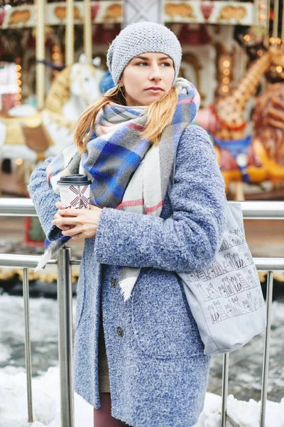 Girl in grey coat, hat and blanket scarf with cup of coffee near carousel — Stock Photo, Image