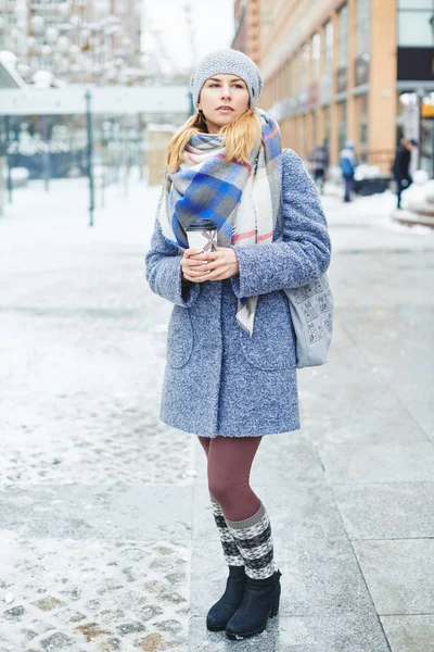 Girl in grey coat, hat and blanket scarf with cup of coffee on winter street — Stock Photo, Image
