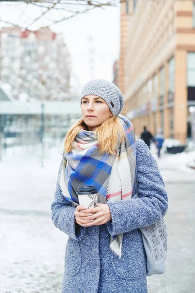 Girl in grey coat, hat and blanket scarf with cup of coffee on winter street — Stock Photo, Image