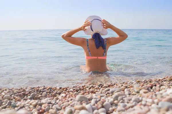 Chica en bikini rosa con sombrero blanco sentado detrás de la cámara en guijarros en la playa, tomando el sol y mirando el mar de aguamarina pura. Perfecta vista de verano concept.slender morena bronceada en traje de baño — Foto de Stock