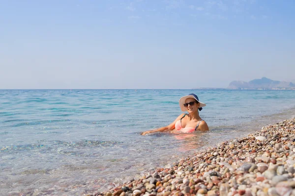 Chica sexy con bikini y sombrero y disfrutar del agua en la playa de guijarros. Mujer hermosa relajarse en la playa de guijarros vacía en el cuerpo natural reserve.well-shaped en bikini sentado en el agua, admirando fascinante — Foto de Stock