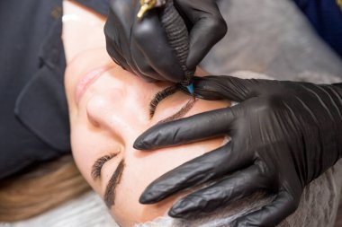 Kalıcı Kaşlar Makyajı. Mikroplu kaş. Yüzüne kaş dövmesi yaptıran bir güzellik uzmanı. Uzun kirpikleri olan genç bir kız güzellik salonunda kaşlarını cımbızla alıyor. Kalıcı makyaj kaşları var, dövme yaptırıyor.