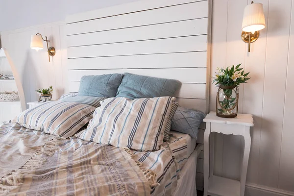 Contemporary Bed dressed with pastel colored cushions and handing lamp.Vase with spring flowers standing on white sidetable.bright bedroom interior. Comfortable bed with coverlet and striped pillows. — Stok fotoğraf