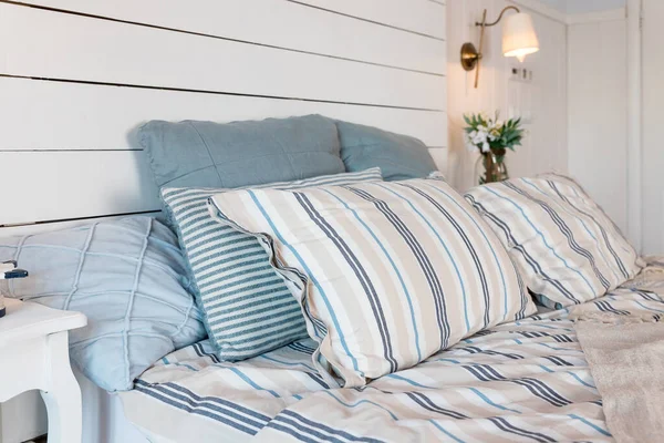 Contemporary Bed dressed with pastel colored cushions and handing lamp.Vase with spring flowers standing on white sidetable.bright bedroom interior. Comfortable bed with coverlet and striped pillows. — Stock Photo, Image