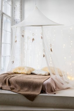 bed with patterned bedding and white drapes in bright bedroom interior.Cozy scandinavian bedroom interior at mansard cabin. Boho style, merino woolen plaid decorated bed, yarn knitted blanket,garland