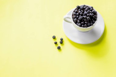 Beyaz kahve fincanında olgun yabanmersini ve sarı arka planda böğürtlen deseni, kopyalama alanı. Bir kaptaki yabanmersinine odaklan. Taze çiğ organik çiftlik yaban mersinleri. Vitaminler, sağlıklı gıda konsepti..