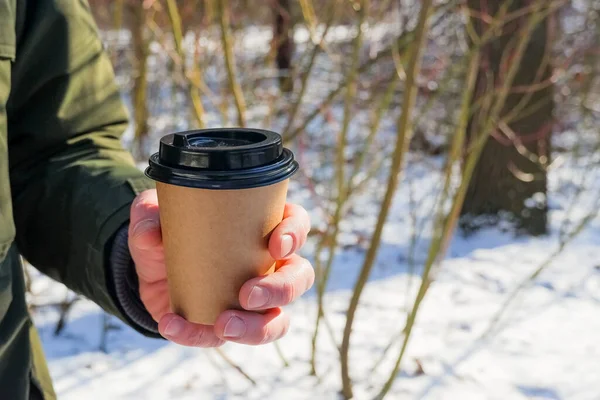 Человек держит чашку кофе в парке со снежным фоном. Coffee to go in winter concept. Горячие напитки зимой скрывают под собой бумажную кружку кофе. Кофе на вынос в холодный зимний день . — стоковое фото