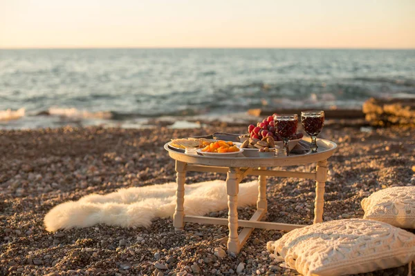 Romantic outdoor dinner with wine and cheese by seaside. Two glasses of wine, grapes, dried fruits and cheese placed on small round table, with white pillows and sheepskin around the table.