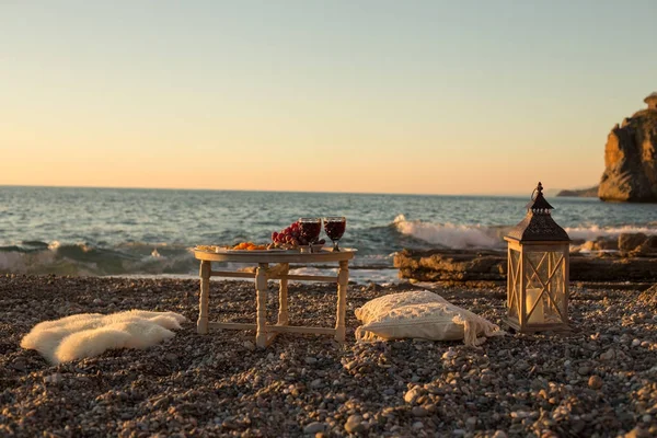 Romantic outdoor dinner with wine and cheese by seaside. Two glasses of wine, grapes, dried fruits and cheese placed on small round table, with white pillows and sheepskin around the table.