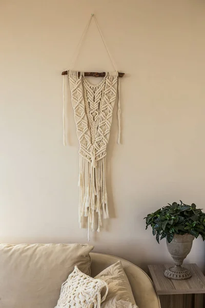 Macrame wall art hanging in the beige interior in beige interior hanging over sofa with green plant in iron vase  placed at he side table