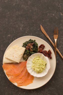 Guacamole, füme somon, spinhach, kapari, kurutulmuş domates ve gözleme ile siyah masa üzerinde sağlıklı kahvaltı tabağı. Dikey kompozisyon. Kopyalama alanı.