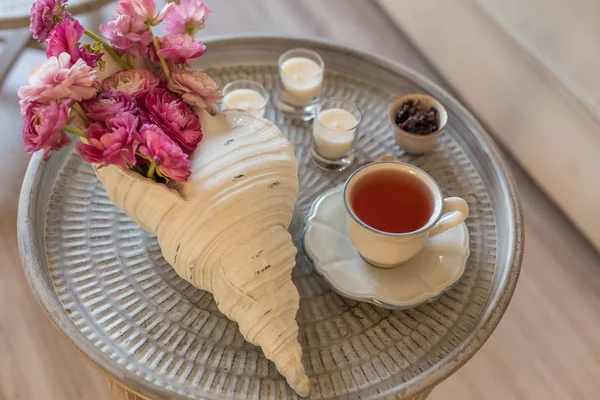 Deniz kabuğu şeklinde vazo pembe ranunculus ile Yuvarlak metal rustik masa, beyaz bir fincan ve üç mum içinde carcade çay