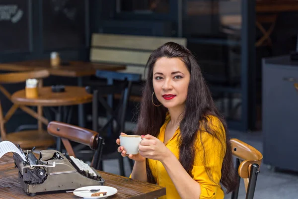 writer female with long dark hair, wearing yellow shirt and brown polka dot skirt, drinking cappuccino in a coffee shop with dark interior and with vintage typewriter on a wooden table