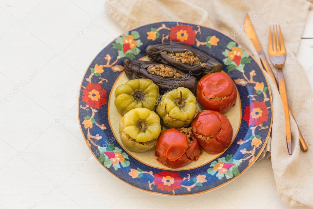 Upper view shot of meat stuffed tomatoes, peppers and eggplant in colorful plate with floral ornament over white wooden background with copper color cutlery laying on cotton linen.