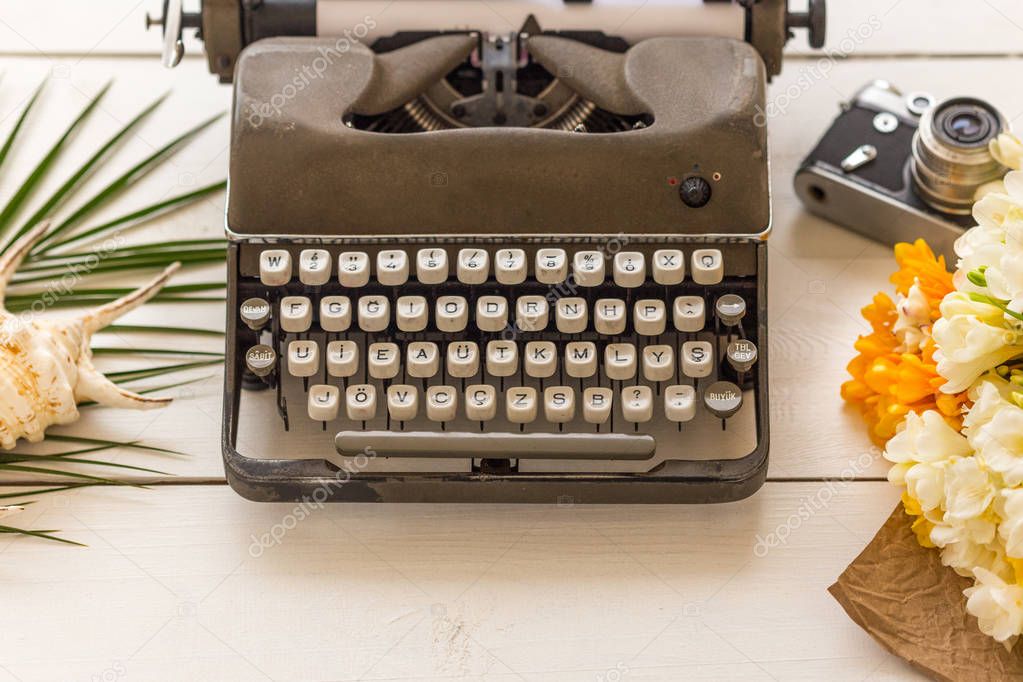 Writing and freelance concept. Retro typewriter with vintage film camera, white and yellow flowers on white wooden tabletop.