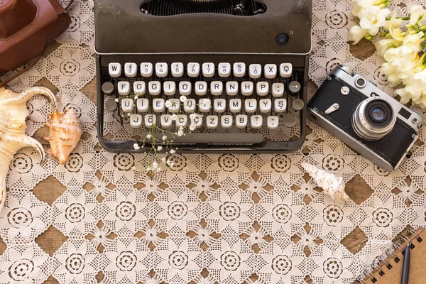 Spring or summer freelance and writing concept. Retro typewriter with gypsophila flower in its keys, vintage film camera and seashells on lace table cover. Top view. Copy space.