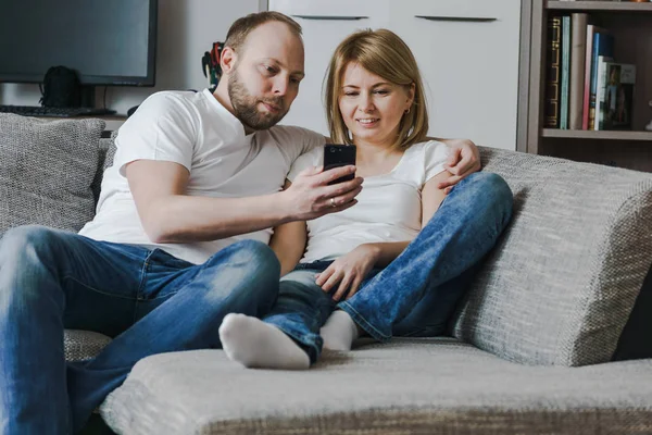 Imagen natural y de estilo de vida de una atractiva pareja sentada en el sofá mirando juntos el teléfono inteligente en la sala de estar . — Foto de Stock