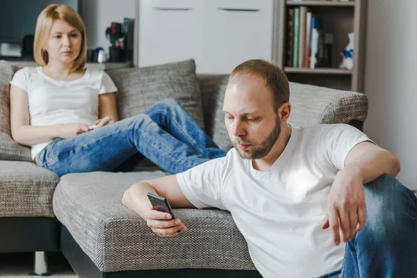 Fiatal nő ül beszél, hogy férje míg ő használja a mobilját a saját nappalijában kanapé. — Stock Fotó