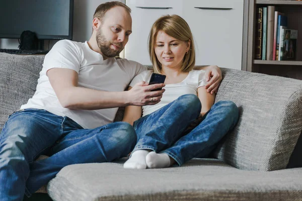 Imagen natural y de estilo de vida de una atractiva pareja sentada en el sofá mirando juntos el teléfono inteligente en la sala de estar . — Foto de Stock