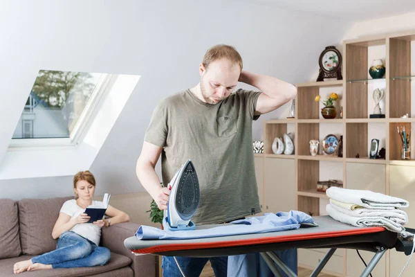 Mujer joven sentada en el sofá leyendo el libro mientras su marido plancha la camisa en casa . — Foto de Stock