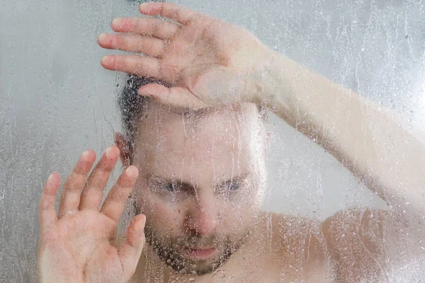 Gestresster Mann duscht unter fließendem Wasser und legt seine Hand auf das Glas in der Duschkabine hinter einer transparenten Nebeltür im Badezimmer — Stockfoto