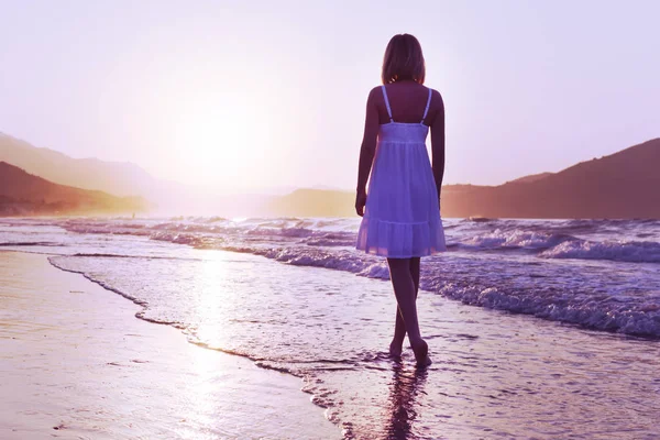 A girl is walking along Crete island coastline at the sunset time. Lilac toning. Chillout relax concept. — Stock Photo, Image