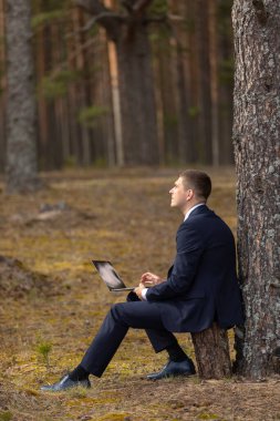 Ormanda dizüstü bilgisayarla dinlenmek. İş adamı elinde dizüstü bilgisayarla parkta oturur ve güneşe bakar. Doğada uzak çalışma