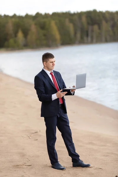 Independiente en la naturaleza. Un hombre trabaja detrás de un portátil en el lago. oficina de empresarios en la playa . — Foto de Stock