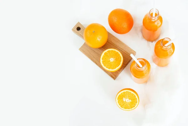 Tres botellas de jugo de naranja y tubos están sobre la mesa sobre un fondo blanco, tanto como dos naranjas y una naranja cortada en tableros de madera . — Foto de Stock
