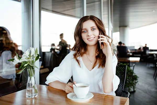 the nice business lady speaks by smartphone in street cafe. the girl is dressed in a white blouse, on a table the cup of coffee stands nearby.