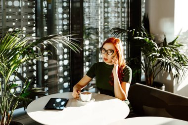 Seksi kırmızı saçlı kız elbise giyen bir gözlük ve yeşil kafede oturur, bir fincan kahve elinde tutan ve telefonla konuşuyor. 