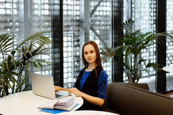 young nice business woman works behind the laptop for little tables in cafe in business center. Nearby there are a cup of coffee and the daily log with records.