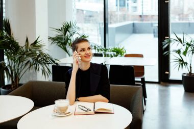 Genç iş kadını ile sıkı bir takım elbise içinde a defter girişleri yapar. Kız ofis kafede oturur ve kahve fincan yakın duruyor.