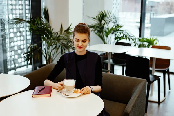 The young business woman in a strict suit makes entries in a notebook. The girl sits in cafe of office and the cup of coffee stands nearby.