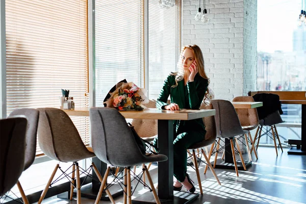 The beautiful young woman in coffee house on an appointment. The blue-eyed blonde in a green suit sits at a little table in cafe and holds phone in hand. Nearby on a table a bouquet of flowers.