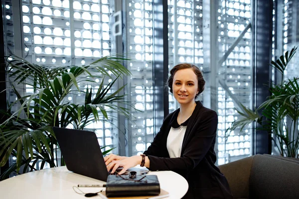 Business portrait. The business woman with collected hair, in a white blouse and a black jacket works behind the laptop at office. Nearby on a table the notebook and office supplies.
