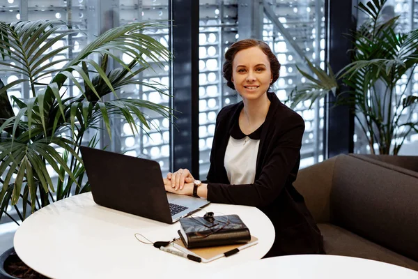 Business portrait. The business woman with collected hair, in a white blouse and a black jacket works behind the laptop at office. Nearby on a table the notebook and office supplies.