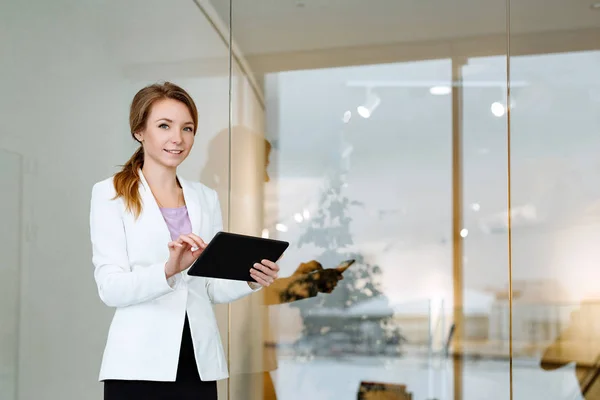 Business a portrait of the positive woman of the blonde in a white jacket against the background of modern office of business center with the black tablet in hands.