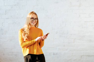Arka planı beyaz duvara sarı bir kazak sarışın genç kız portresi. Kız gülümsüyor ve görüşmeler smartphone. Kopya alanı.