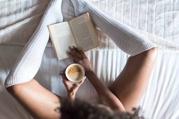 Γυναίκα διαβάζοντας ένα βιβλίο και να πίνει καφέ στο κρεβάτι με κάλτσες — Φωτογραφία Αρχείου