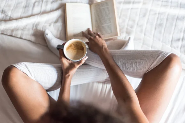 Μαύρη γυναίκα διαβάζοντας ένα βιβλίο και να πίνει καφέ στο κρεβάτι με κάλτσες — Φωτογραφία Αρχείου