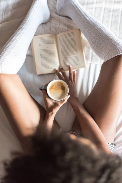 Чорна жінка читає книгу і п'є каву на ліжку з шкарпетками — стокове фото