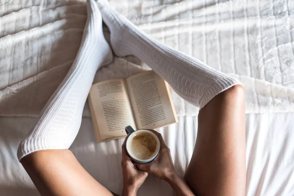 Woman reading a book and drinking coffee on bed with socks — Stock Photo, Image