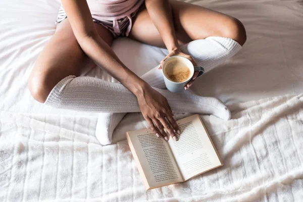 Woman reading a book and drinking coffee on bed with socks — Stock Photo, Image