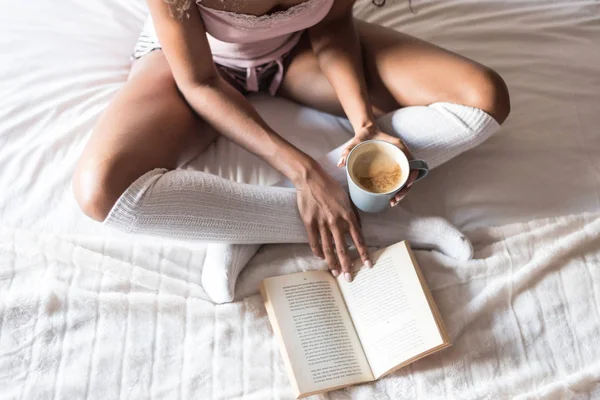 Μαύρη γυναίκα διαβάζοντας ένα βιβλίο και να πίνει καφέ στο κρεβάτι με κάλτσες — Φωτογραφία Αρχείου