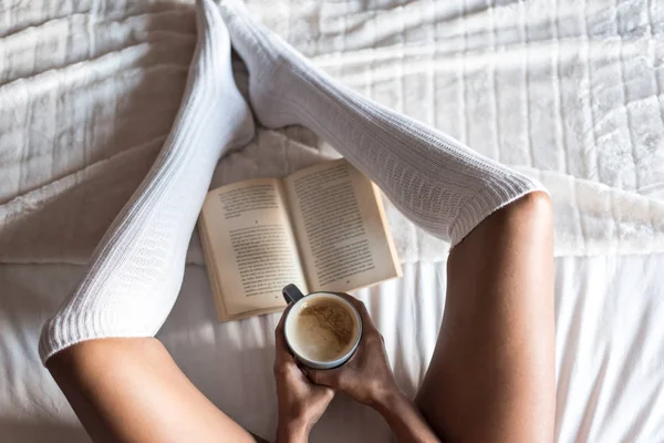 Donna che legge un libro e beve caffè sul letto con le calze — Foto Stock