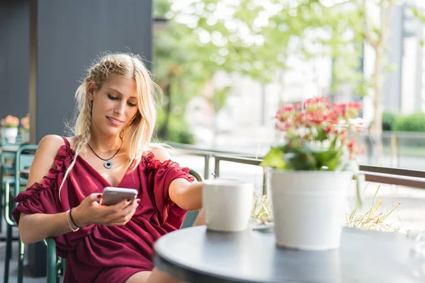 Feliz rubia hermosa mujer usando un teléfono móvil bebiendo una taza de café en una terraza al aire libre — Foto de Stock
