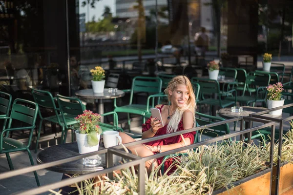 Happy blonde beautiful woman using a mobile phone drinking a cup of coffee in a terrace outdoors — Stock Photo, Image