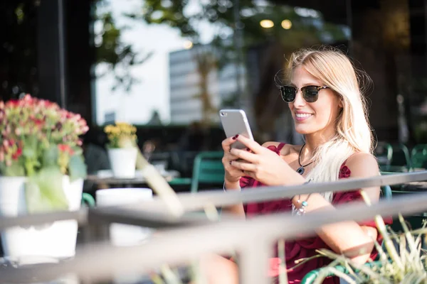 Feliz rubia hermosa mujer usando un teléfono móvil bebiendo una taza de café en una terraza al aire libre — Foto de Stock