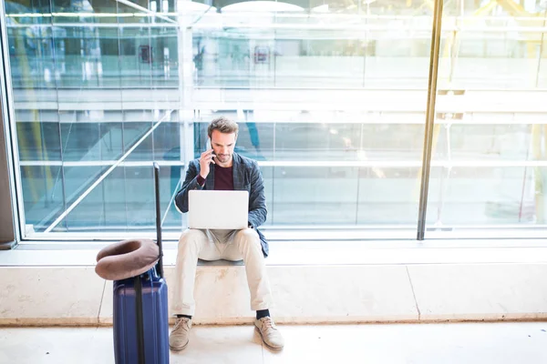 Hombre sentado en el aeropuerto usando el ordenador portátil y el teléfono móvil al lado de — Foto de Stock